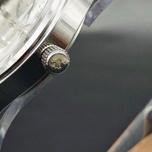 May include: Close-up of a silver wristwatch. The watch features a silver case, a textured crown with a logo, and a clear watch face. The watch is set against a dark background, highlighting its metallic sheen.
