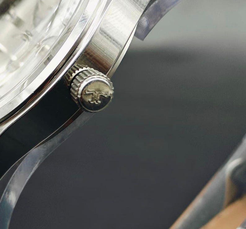 May include: Close-up of a silver wristwatch. The watch features a silver case, a textured crown with a logo, and a clear watch face. The watch is set against a dark background, highlighting its metallic sheen.
