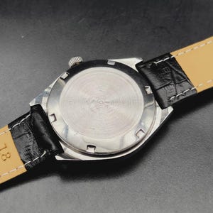 May include: A silver-toned wristwatch case with a circular brushed metal back featuring six notches around the perimeter. The watch is attached to a black leather strap with a textured crocodile-like pattern and white stitching along the edges.