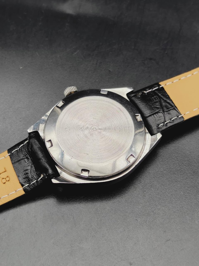 May include: A silver-toned wristwatch case with a circular brushed metal back featuring six notches around the perimeter. The watch is attached to a black leather strap with a textured crocodile-like pattern and white stitching along the edges.