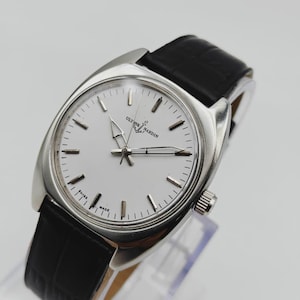 May include: A silver-toned Ulysse Nardin wristwatch with a white face and black leather band. The watch features a round face with silver-toned hands and markers. The band is black, and the watch is displayed on a clear stand.