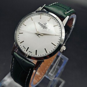 Pu&ograve; includere: Un orologio da polso argentato con quadrante bianco e indicatori neri. L'orologio presenta la scritta "JAEGER-LECOULTRE CLUB SWISS MADE" e ha un cinturino in pelle verde con cuciture bianche. L'orologio &egrave; esposto su un supporto trasparente.