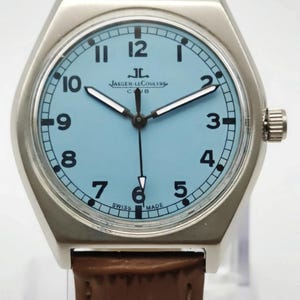 K&ouml;nnte beinhalten: Eine silberfarbene Armbanduhr mit hellblauem Zifferblatt und braunem Lederarmband. Das Zifferblatt zeigt schwarze Ziffern und den Text "JAEGER-LECOULTRE CLUB SWISS MADE". Die Uhr hat eine silberfarbene L&uuml;nette und eine Krone auf der rechten Seite.