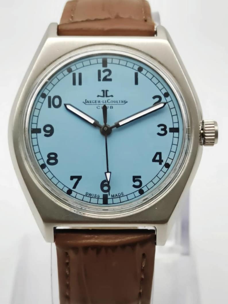K&ouml;nnte beinhalten: Eine silberfarbene Armbanduhr mit hellblauem Zifferblatt und braunem Lederarmband. Das Zifferblatt zeigt schwarze Ziffern und den Text "JAEGER-LECOULTRE CLUB SWISS MADE". Die Uhr hat eine silberfarbene L&uuml;nette und eine Krone auf der rechten Seite.