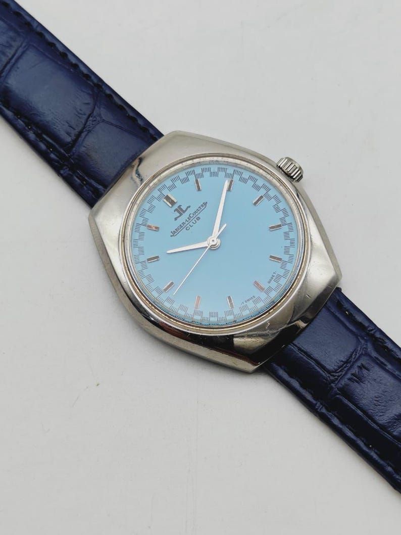 May include: A silver Jaeger-LeCoultre Club watch with a light blue face and a dark blue leather strap. The watch face has silver hands and hour markers. The strap is approximately 20 cm long.