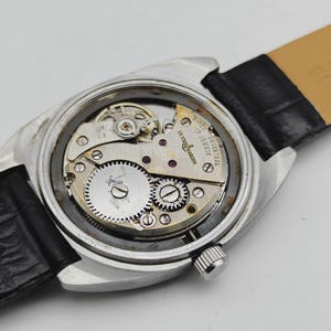 May include: Close-up of a vintage Ulysse Nardin watch, showcasing the intricate mechanical movement. The watch has a silver-toned case, a black leather strap, and visible gears and jewels. The text "Ulysse Nardin 17 Jewels" is visible on the watch face.