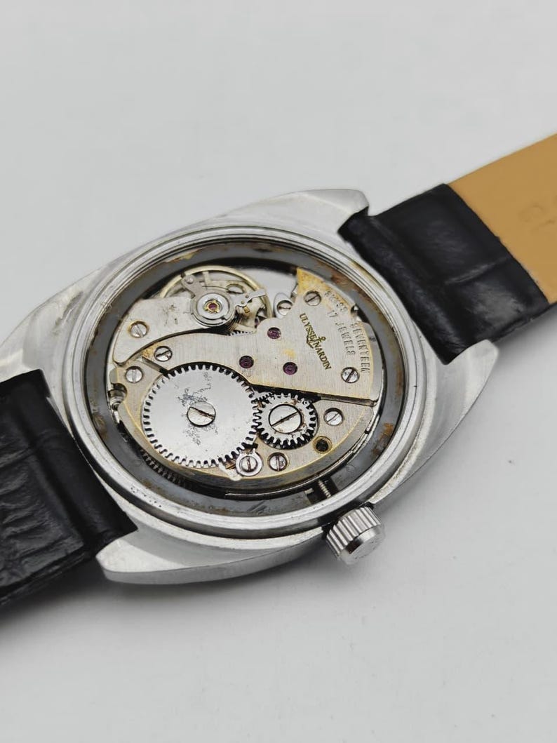 May include: Close-up of a vintage Ulysse Nardin watch, showcasing the intricate mechanical movement. The watch has a silver-toned case, a black leather strap, and visible gears and jewels. The text "Ulysse Nardin 17 Jewels" is visible on the watch face.