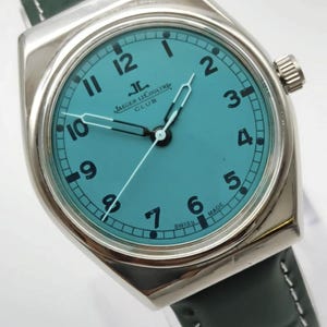 Pode incluir: Um rel&oacute;gio prateado com mostrador azul-turquesa e algarismos pretos. O rel&oacute;gio apresenta o texto "JAEGER LECOULTRE CLUB" e "SWISS MADE". Possui uma pulseira de couro verde escuro.
