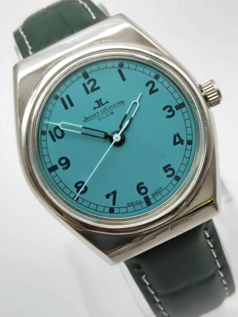 Pode incluir: Um rel&oacute;gio prateado com mostrador azul-turquesa e algarismos pretos. O rel&oacute;gio apresenta o texto "JAEGER LECOULTRE CLUB" e "SWISS MADE". Possui uma pulseira de couro verde escuro.