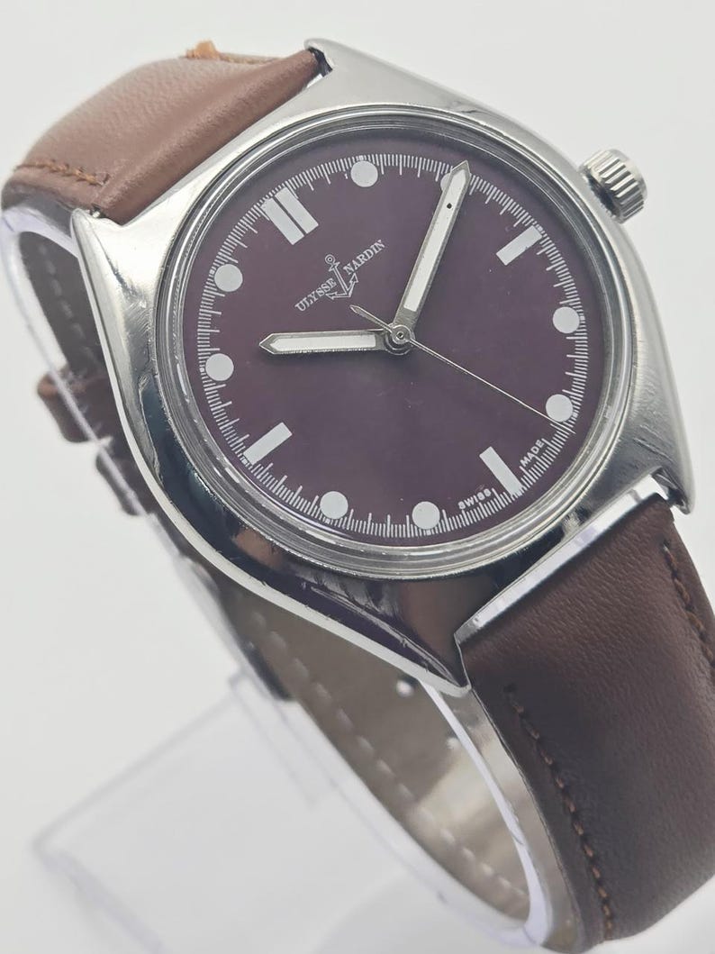 May include: A silver-toned wristwatch with a dark burgundy face and a brown leather band. The watch face features white hour markers and hands, with the brand name "Ulysse Nardin" displayed. The watch has a classic, elegant design.