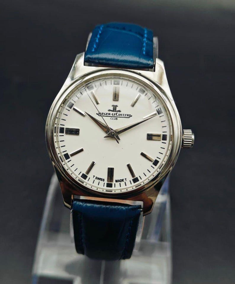 Peut inclure: Une montre Jaeger-LeCoultre Club argent&eacute;e avec un cadran blanc et un bracelet en cuir bleu. Le cadran comporte des marqueurs et des aiguilles noirs, avec le nom de la marque et le texte "T SWISS MADE T". Le bracelet est bleu vif.