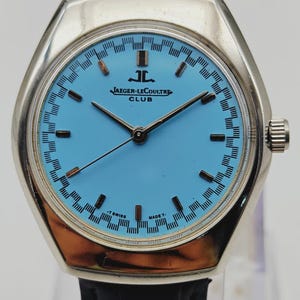 May include: A vintage Jaeger-LeCoultre Club watch with a light blue face and silver case. The watch has black hands and hour markers, and a blue leather strap. The watch face displays the Jaeger-LeCoultre logo and the word "CLUB".