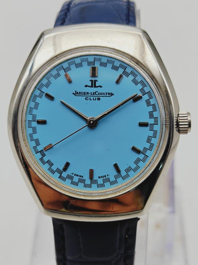 May include: A vintage Jaeger-LeCoultre Club watch with a light blue face and silver case. The watch has black hands and hour markers, and a blue leather strap. The watch face displays the Jaeger-LeCoultre logo and the word "CLUB".