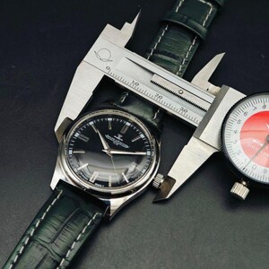 K&ouml;nnte beinhalten: Eine silberne Armbanduhr mit schwarzem Zifferblatt und dunkelgr&uuml;nem Lederarmband wird mit einem Messschieber gemessen. Die Uhr hat silberne Zeiger und Markierungen. Das Armband ist mit wei&szlig;em Faden gen&auml;ht. Eine Messuhr ist ebenfalls sichtbar.