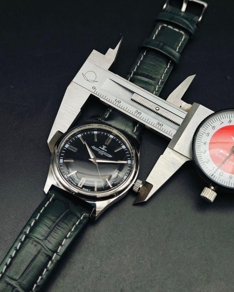 K&ouml;nnte beinhalten: Eine silberne Armbanduhr mit schwarzem Zifferblatt und dunkelgr&uuml;nem Lederarmband wird mit einem Messschieber gemessen. Die Uhr hat silberne Zeiger und Markierungen. Das Armband ist mit wei&szlig;em Faden gen&auml;ht. Eine Messuhr ist ebenfalls sichtbar.