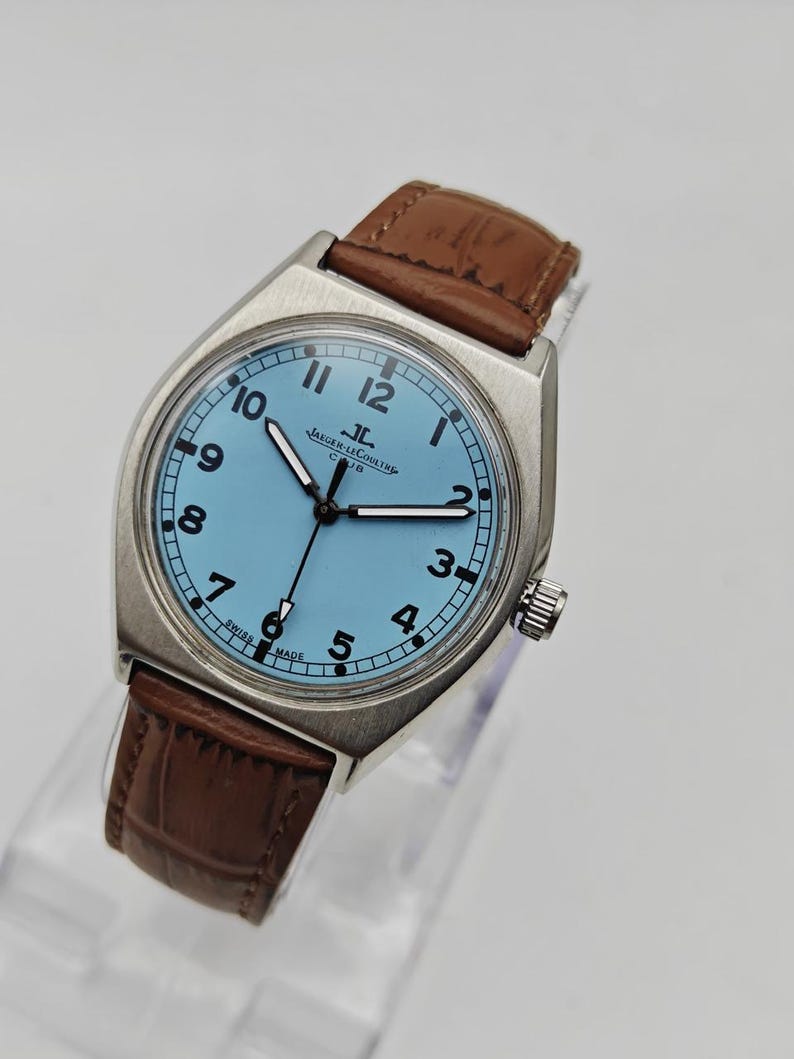K&ouml;nnte beinhalten: Eine silberfarbene Armbanduhr mit hellblauem Zifferblatt und braunem Lederarmband. Das Zifferblatt zeigt schwarze Ziffern und Zeiger, mit dem Markennamen "JAEGER-LECOULTRE". Die Uhr hat ein klassisches Design.