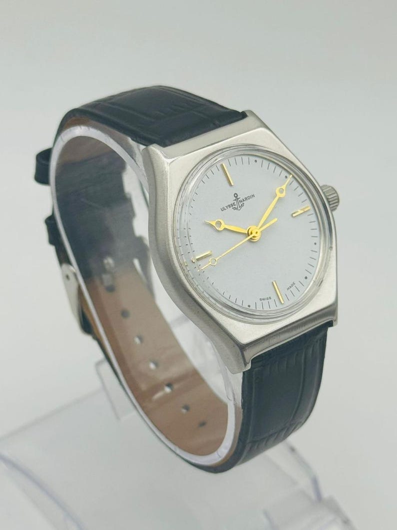 May include: A silver-toned Ulysse Nardin watch with a white face, gold hands, and a black leather band. The watch has a rectangular case and a small crown. The watch face features the brand name and a nautical symbol.