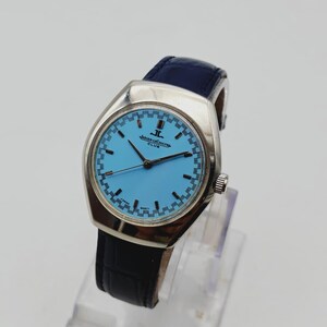 May include: A vintage Jaeger-LeCoultre Club watch with a light blue face and a silver case. The watch has a dark blue leather strap and a silver crown. The watch is displayed on a clear stand.
