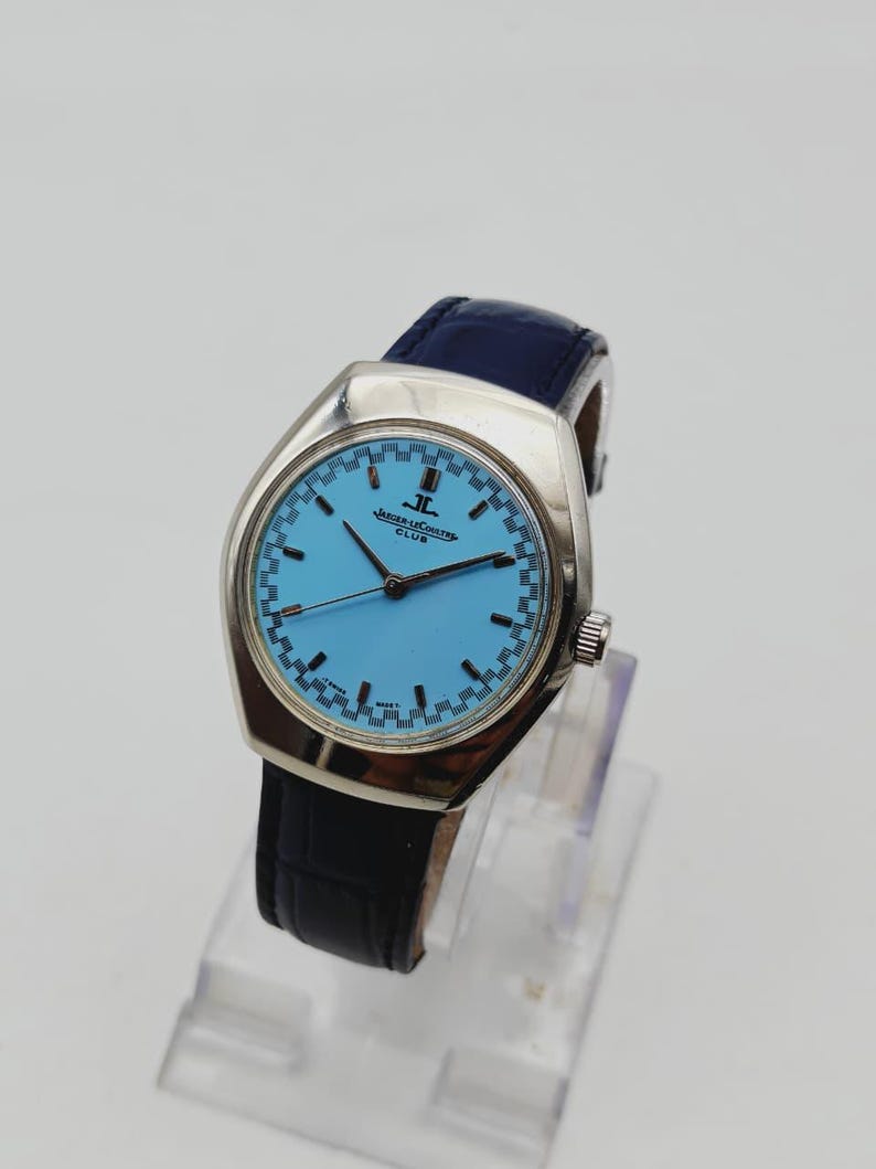 May include: A vintage Jaeger-LeCoultre Club watch with a light blue face and a silver case. The watch has a dark blue leather strap and a silver crown. The watch is displayed on a clear stand.