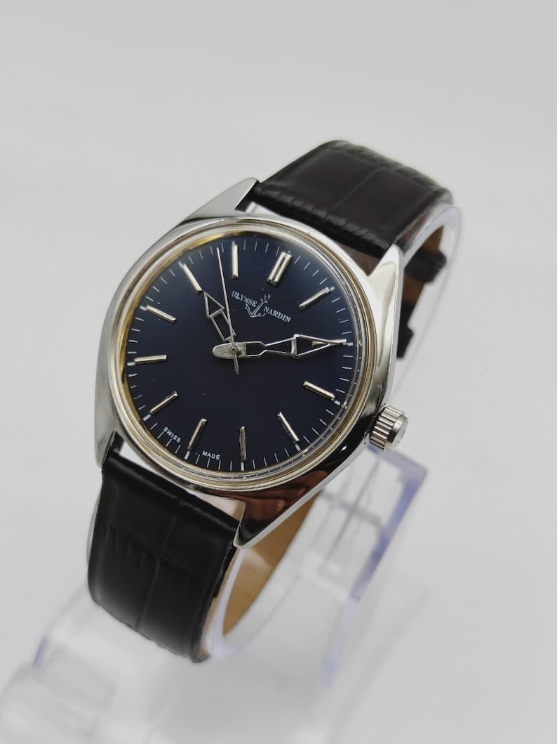 May include: A silver-toned Ulysse Nardin wristwatch with a dark blue face and black leather band. The watch features silver hands and markers, and the text "Ulysse Nardin Swiss Made" is visible on the face. The band has a crocodile pattern.