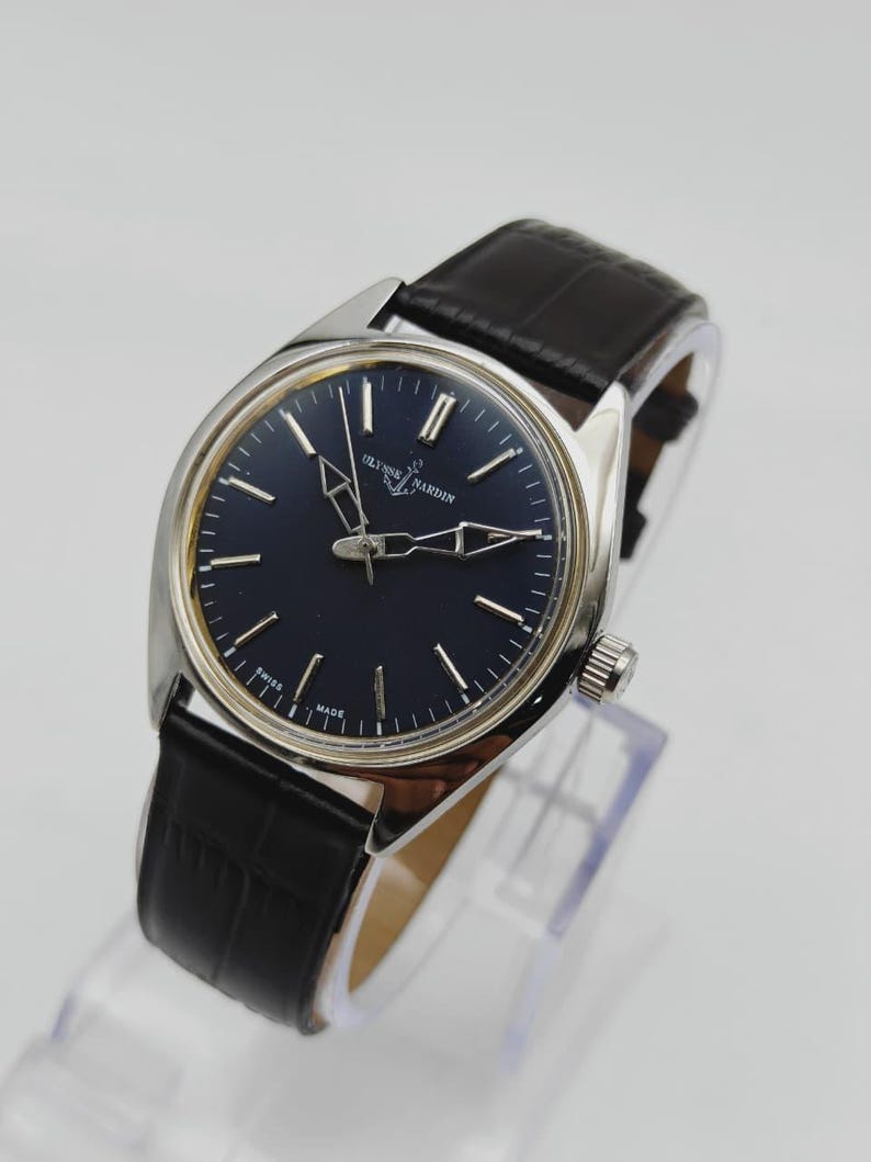 May include: A silver-toned Ulysse Nardin wristwatch with a dark blue face and black leather band. The watch features silver hands and markers, and the band has a textured, crocodile-like pattern. The watch is displayed on a clear stand.