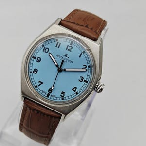 K&ouml;nnte beinhalten: Eine silberfarbene Armbanduhr mit hellblauem Zifferblatt und braunem Lederarmband. Das Zifferblatt zeigt schwarze Ziffern und Zeiger, mit dem Markennamen "JAEGER-LECOULTRE". Die Uhr hat ein klassisches Design.