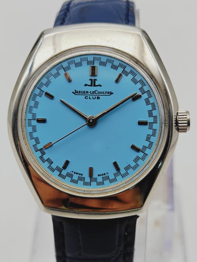 May include: A vintage Jaeger-LeCoultre Club watch with a light blue face and silver case. The watch has black hands and hour markers, and a blue leather strap. The watch face displays the Jaeger-LeCoultre logo and the word "CLUB".