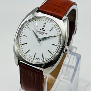 May include: A silver-toned Jaeger-LeCoultre Club watch with a white face, black hour markers, and green accents. The watch has a brown leather strap. The watch face displays the Jaeger-LeCoultre logo and the words "CLUB".