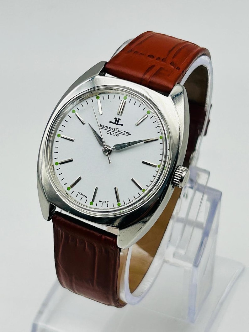 May include: A silver-toned Jaeger-LeCoultre Club watch with a white face, black hour markers, and green accents. The watch has a brown leather strap. The watch face displays the Jaeger-LeCoultre logo and the words "CLUB".