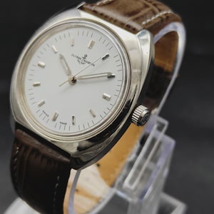 May include: A silver-tone Ulysse Nardin wristwatch featuring a round white dial with silver hour markers, silver hands, and a brown leather strap with a textured finish. The watch face displays the brand name and an anchor logo at the top.