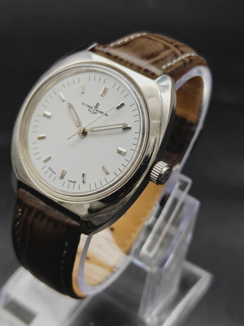 May include: A silver-tone Ulysse Nardin wristwatch featuring a round white dial with silver hour markers, silver hands, and a brown leather strap with a textured finish. The watch face displays the brand name and an anchor logo at the top.