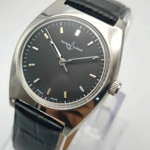 Puede incluir: Un reloj de pulsera plateado con esfera negra y correa de cuero negro. La esfera del reloj muestra la marca "ULYSSE NARDIN" e indicadores horarios dorados. El reloj est&aacute; marcado "SWISS MADE".