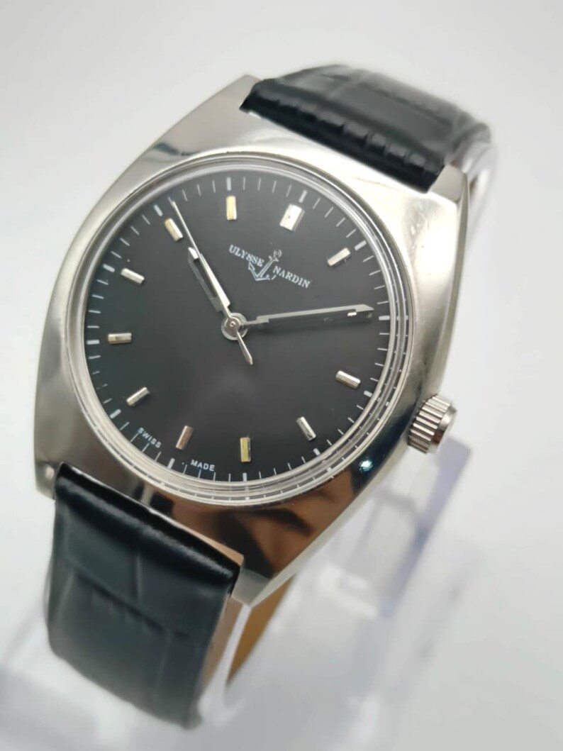 Puede incluir: Un reloj de pulsera plateado con esfera negra y correa de cuero negro. La esfera del reloj muestra la marca "ULYSSE NARDIN" e indicadores horarios dorados. El reloj est&aacute; marcado "SWISS MADE".