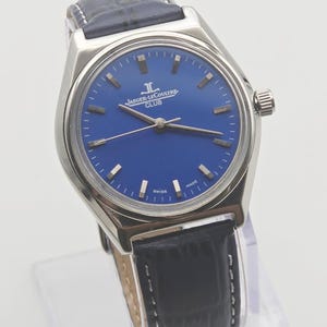 May include: A stainless steel Jaeger-LeCoultre Club watch with a blue dial and black leather strap. The watch features silver hour markers and hands. The dial has the Jaeger-LeCoultre logo and the word "CLUB" printed on it. The watch is marked "SWISS MADE".