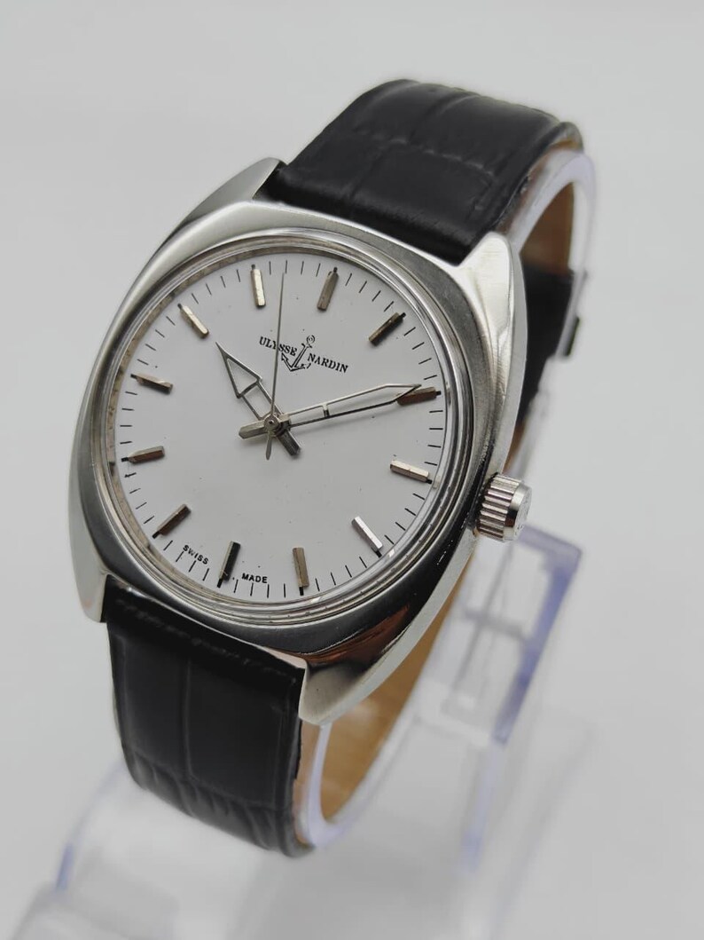 May include: A silver-toned wristwatch with a white face and black leather band. The watch face features black hour markers and the brand name "Ulysse Nardin". The watch is displayed on a clear stand.
