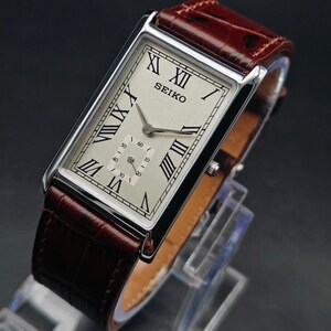 May include: A rectangular Seiko wristwatch with a silver-colored face, black Roman numerals, and a subdial. The watch has a brown leather band. The watch is displayed on a clear stand against a dark background.