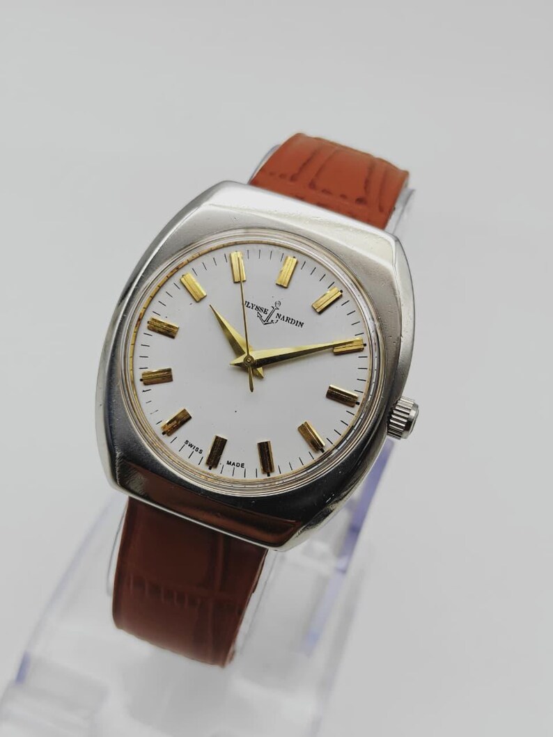 May include: A vintage Ulysse Nardin wristwatch with a silver-toned case and a white dial. The watch features gold-toned hour markers and hands, and a brown leather strap. The watch is marked "Swiss Made".