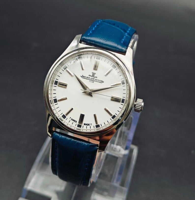 Peut inclure: Une montre-bracelet argent&eacute;e avec un cadran blanc et un bracelet en cuir bleu. Le cadran comporte des marqueurs et des aiguilles noirs, avec la marque "Jaeger-LeCoultre Club" visible. Le bracelet est bleu vif.