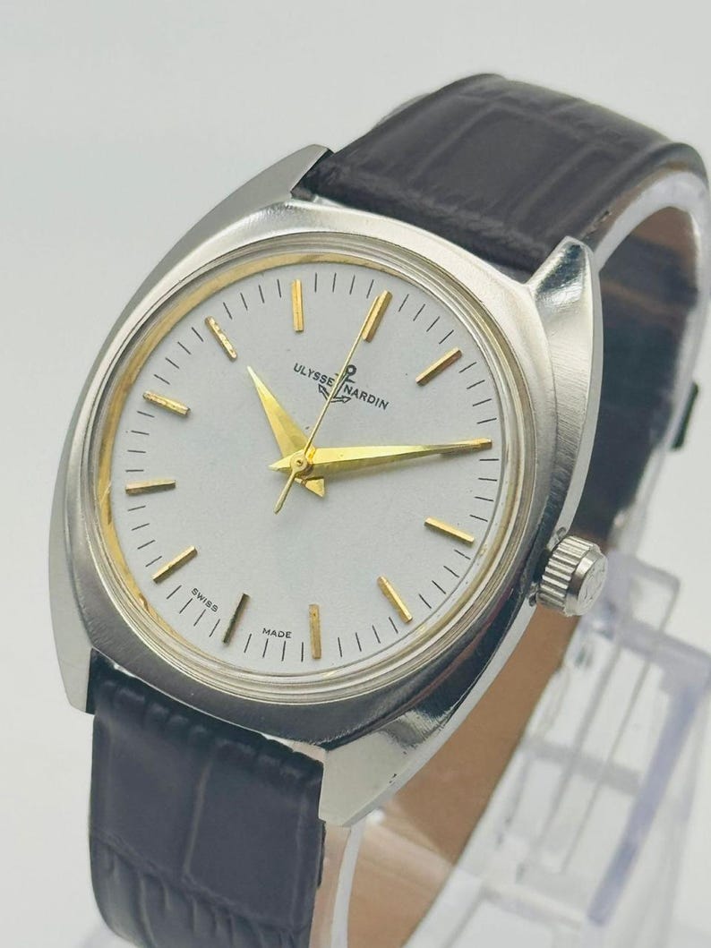 May include: A vintage Ulysse Nardin wristwatch with a silver case and a white face. The watch has gold hands and markers, and a black leather strap. The watch is marked "Swiss Made".