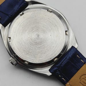 May include: A silver wristwatch with a blue leather strap. The watch's back casing is visible, with a circular, textured pattern. The blue strap is stitched to a tan underside. The watch crown is visible on the side.