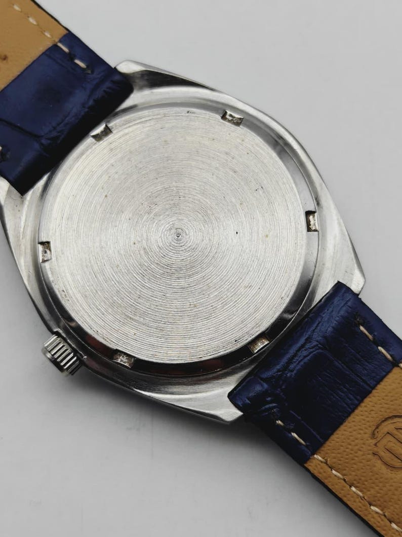 May include: A silver wristwatch with a blue leather strap. The watch's back casing is visible, with a circular, textured pattern. The blue strap is stitched to a tan underside. The watch crown is visible on the side.