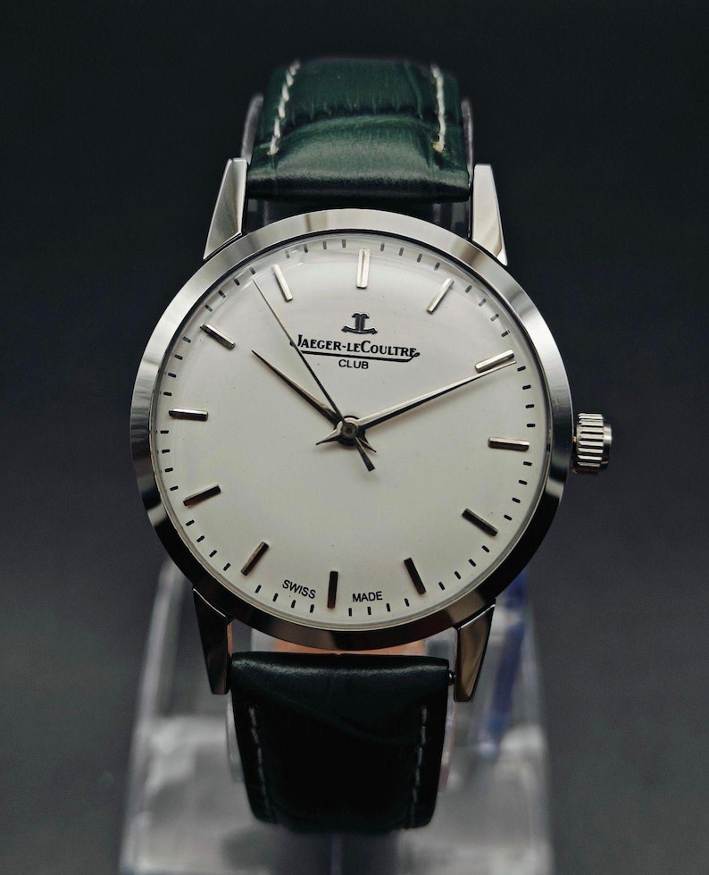 Pu&ograve; includere: Un orologio Jaeger-LeCoultre Club argentato con quadrante bianco, lancette nere e cinturino in pelle nera. Il quadrante mostra il logo Jaeger-LeCoultre e le parole "SWISS MADE". L'orologio ha un cinturino in pelle verde.
