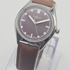 May include: A silver-toned wristwatch with a brown face and a brown leather band. The watch face has white markers and hands, with the brand name "ULYSSE NARDIN" displayed. The watch is displayed on a clear stand.