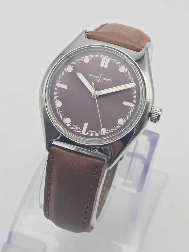 May include: A silver-toned wristwatch with a brown face and a brown leather band. The watch face has white markers and hands, with the brand name "ULYSSE NARDIN" displayed. The watch is displayed on a clear stand.