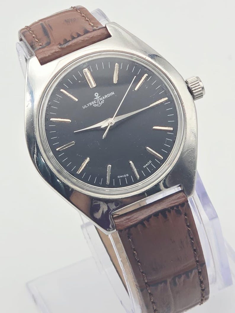 May include: A silver-toned Ulysse Nardin wristwatch with a black face and brown leather band. The watch features silver hands and markers, and the brand's logo is visible. The watch is displayed on a clear stand.