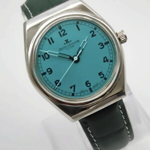 Pode incluir: Um rel&oacute;gio Jaeger-LeCoultre Club prateado com mostrador turquesa e ponteiros pretos. O rel&oacute;gio tem algarismos pretos e uma pulseira de couro preta com costuras brancas. O mostrador diz "JAEGER LECOULTRE CLUB" e "SWISS MADE".
