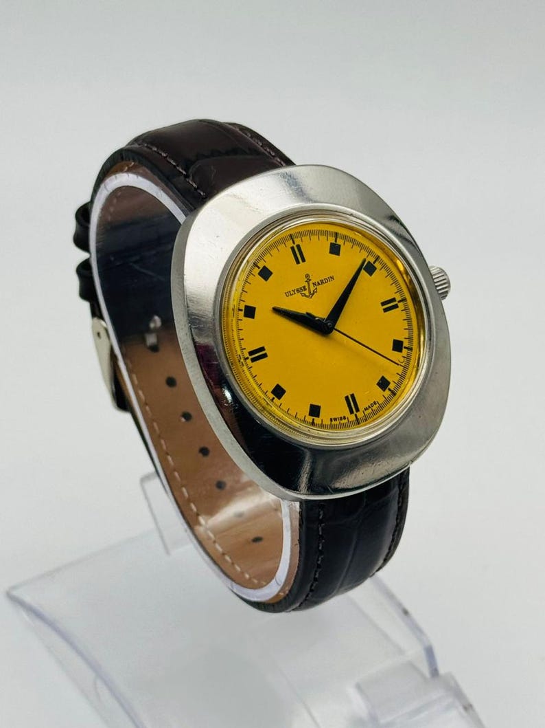 May include: A vintage Ulysse Nardin watch with a bright yellow face and black hour markers. The watch has a silver-tone case and a dark brown leather strap. The watch is displayed on a clear stand.