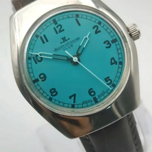 Op de afbeelding: Een zilverkleurig polshorloge met een turquoise wijzerplaat en zwarte cijfers. Het horloge heeft een bruine leren band en een zilveren lunette. De wijzerplaat toont het Jaeger-LeCoultre Club logo. De wijzers zijn zilver.
