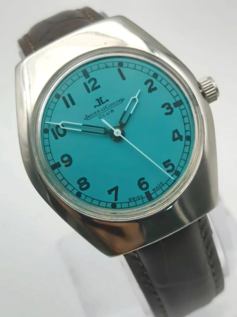 Op de afbeelding: Een zilverkleurig polshorloge met een turquoise wijzerplaat en zwarte cijfers. Het horloge heeft een bruine leren band en een zilveren lunette. De wijzerplaat toont het Jaeger-LeCoultre Club logo. De wijzers zijn zilver.