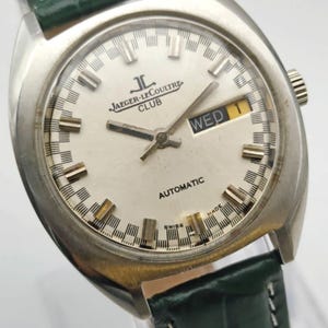 Puede incluir: Un reloj autom&aacute;tico Jaeger-LeCoultre Club vintage con esfera plateada, visualizaci&oacute;n de d&iacute;a y fecha y correa de cuero verde. El reloj tiene una caja de acero inoxidable y presenta el texto "Jaeger-LeCoultre Club" y "Automatic".